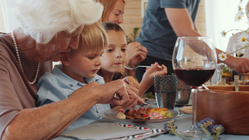 Loving grandmother using knife and fork while feeding cute little grandson at home holiday dinner with large family Royalty-Free Stock Footage #1060664476