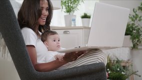 Smiling mixed race woman video conference with friends or relatives holding baby in her arms and waving affably at laptop camera