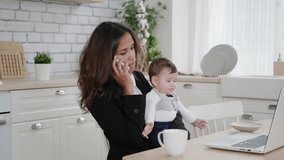 Successful mixed race business woman holds a small child in her arms, communicates with colleagues on a mobile phone and works on a laptop online remotely.