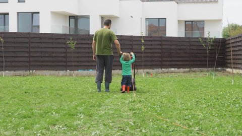 Father and son with back, wearing rubber boots, hold together lawn mower work in garden, kid lean, determined and diligent little child