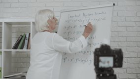 Online female teacher conducts webinars, classes or school lessons. A senior woman stands near flip chart and explains something to the camera