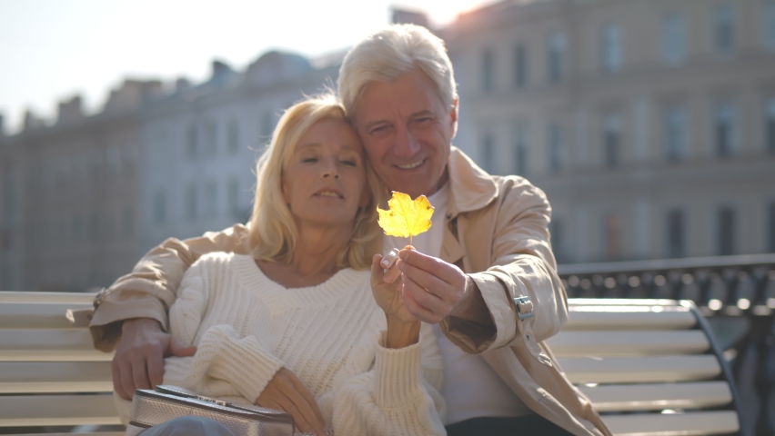 Elegant elderly couple relaxing on bench in city and holding yellow tree leaf