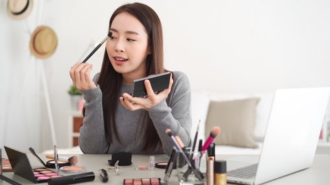 Young beautiful Asian woman professional beauty vlogger influencer doing cosmetic makeup tutorial live streaming with mobile phone at home
