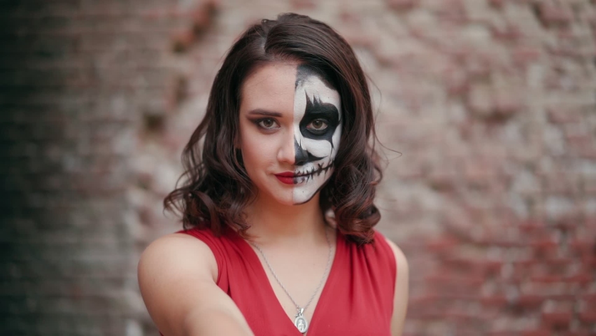 Portrait beautiful woman in a red evening dress and awesome Halloween makeup lures to itself against the backdrop of an abandoned building. Greasepaint for the Holiday on October 31. Slow motion.