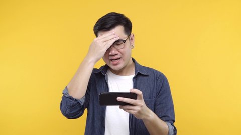 A man put on glasses and denim jacket white t-shirt is playing game on smartphone using modern technology - apps, social networks feel sad isolated on yellow background. 4k resolution.