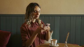 Beautiful blonde woman taking pictures of a cup of coffee with her smartphone and texting afterwards. Young food blogger with cell phone in a coffee shop. Communication, social network concept.