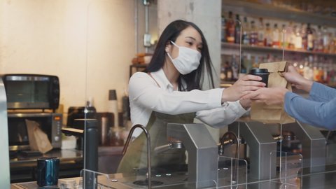 Cheerful asian waitress in white uniform delivering brown empty blank paper bag between acrylic partition to customer at counter in restaurant and both wearing face mask. Pandemic coronavirus 2019.