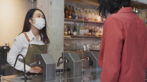 Cheerful asian waitress in white uniform receive food and drink order from caucasian customer at counter behind acrylic partition in restaurant and both wearing face mask. Pandemic coronavirus 2019.