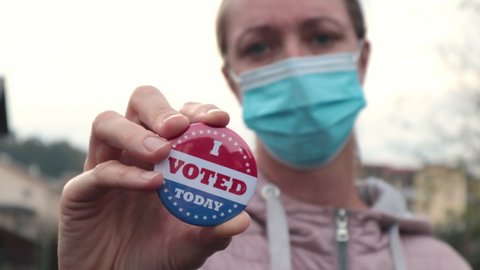Woman with face mask showing I voted today button at american elections outside.