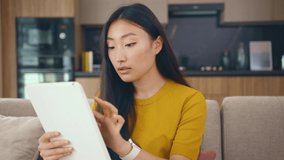Face young asian woman sitting on couch in cozy room use tablet device smile at home. Businesswoman internet confident chatting. Portrait. Slow motion