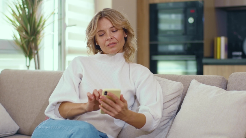 Happy young blond woman use phone social media platform and sit on sofa at home. Internet couch beautiful message indoor apartment. Close up. Slow motion | Shutterstock HD Video #1060711693