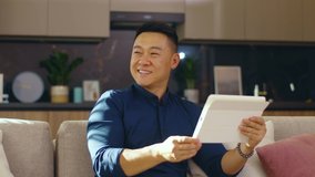 Smiling asian young man use tablet computer sitting on sofa at home. Feel happy. Chinese businessman device handsome. Communication. Close up. Slow motion - Powered by Shutterstock - Get 15% off with code: PIKWIZARD15