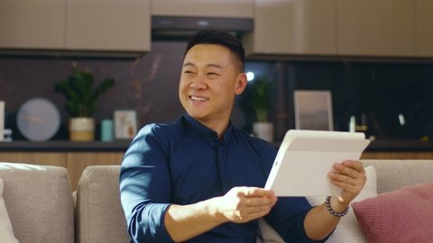 Smiling asian young man use tablet computer sitting on sofa at home. Feel happy. Chinese businessman device handsome. Communication. Close up. Slow motion
