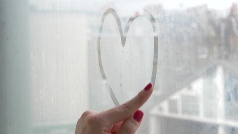 Woman drawing heart shape on the window with her finger - alone young woman draw on wet glass of the window on a moody day isolating at home