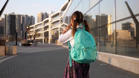 Little child girl with a backpack in school uniform is going to school in city landscape, rear view. Primary school pupil is going to study, steadicam shot.