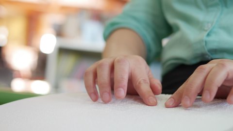 Close-up of disability blind person woman hands moving fingers reading Braille book studying in creative library. Braille is a system of raised dots that can be read with the fingers