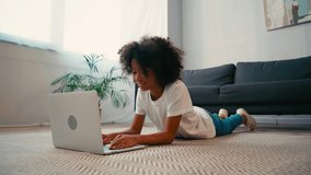 African american girl typing on laptop while lying on carpet at home