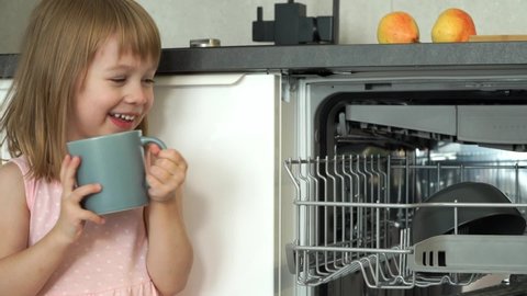 Loading dishwasher. Little girl smiles and laughs, puts cup in dishwasher. Happy child helps with housework. Household and dishwashing concept. Close up.