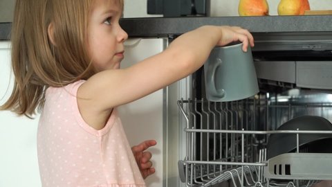 Little girl takes cup out of dishwasher, smiles and laughs. Happy child helps with housework. Unloading dishwasher. Close up
