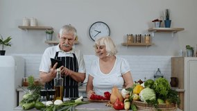 Vegan senior couple cooking salad with raw vegetables while looking on screen of digital tablet for online recipe. Funny mature woman dancing holding fresh red slices of pepper on eyes. 6k downscale
