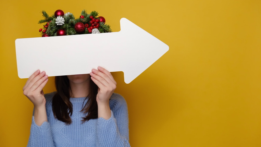 Stunned emotive young woman hides face behind white paper arrow and points sideways, wears Christmas wreath, stands against yellow background with copy space. Happy New Year celebration merry holiday