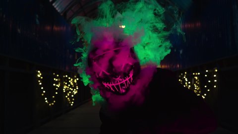 The man in the neon mask blows smoke. Thick green and pink smoke. Scary Halloween Purge killer costume.