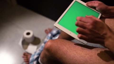 Man hold tablet in hands and swiping to right. Green screen on device or gadget. Guy sit on toilet in rest room. Zoom in picture. Toilet paper on floor.