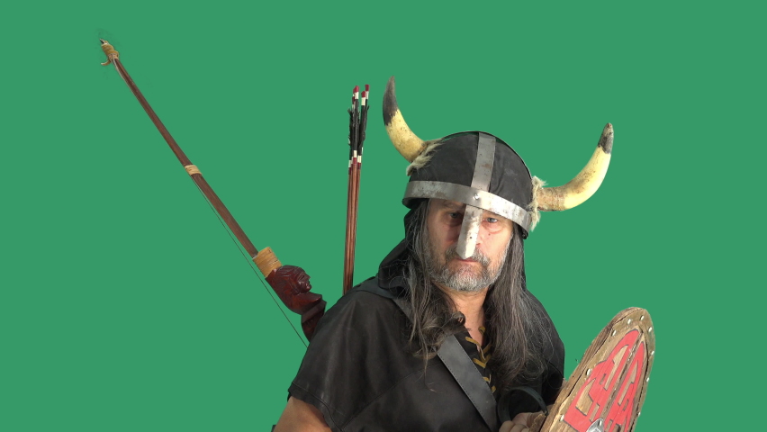 Portrait of formidable commoner warrior against green background, raises silver horn with mead and drinks. Mature man in helmet with horns and with wooden shield