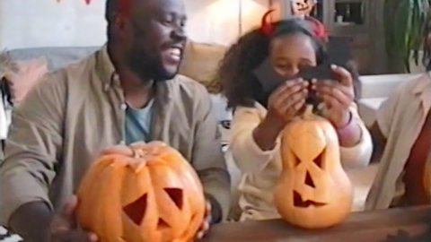 Retro VHS footage of happy African American family playing with devil horns headband, waving at camera and decorating jack-o-lanterns with paper bat on Halloween at home