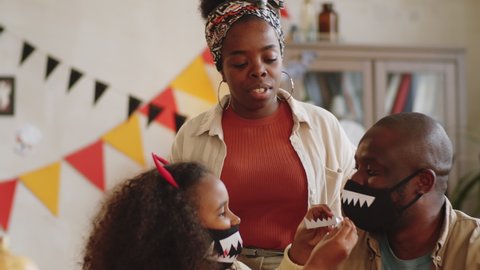 Little African American girl attaching vampire teeth sticker on black face mask of father with help of mom while preparing Halloween costumes for family celebration at home