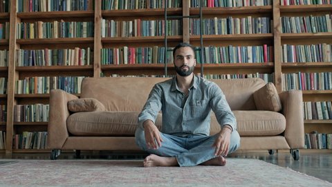 Young Man Meditates In The Library. Guy Is Sitting On The Floor Near The Sofa With His Eyes Closed. He Has Headphones In His Ears.
