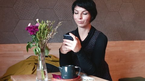 A lonely woman sits at a table in an indoor cafe looking at the phone screen. Addiction to online never ending level game played with friends. The mood improves when the next level is passed.