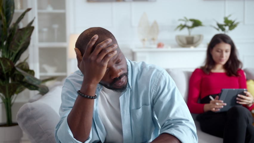 Pensive young african sad man staying in living room with his wife working remotely on tablet computer. Internet addiction. Marriage crisis. Relationships. | Shutterstock HD Video #1060734700