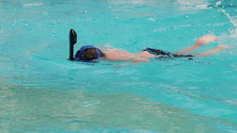Unrecognizable boy in an underwater mask with a snorkel floats on the blue surface of the water in slow motion in a resort pool