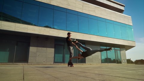 Couple of acrobats on roller skates doing extreme stunts in urban setting. Arc shot of man holding woman by hand and foot while spinning around and she doind tricks. Concept of sport