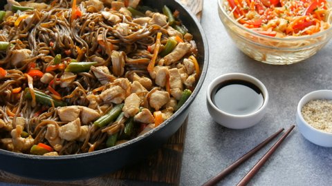 Japanese soba rice noodles with pieces of chicken, carrot, green shoots, sesame seeds, soy sauce. National food in a restaurant in large frying pan next to wooden bamboo sticks. Glass noodle salad.