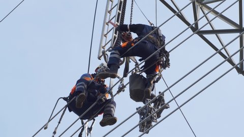 Two utility workers in hardhats sit on electrical high voltage power lines for repair