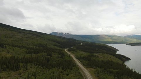 Beautiful View of Road alongside Scenic Lake surrounded by Mountains and Trees on a Cloudy Day. Aerial Drone Shot. Taken near Klondike Highway, Southern Yukon, Canada.