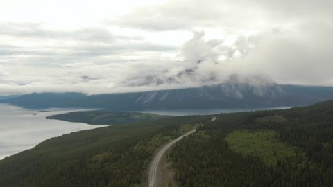 Beautiful View of Road alongside Scenic Lake surrounded by Mountains and Trees on a Cloudy Day. Aerial Drone Shot. Taken near Klondike Highway, Southern Yukon, Canada. 4K