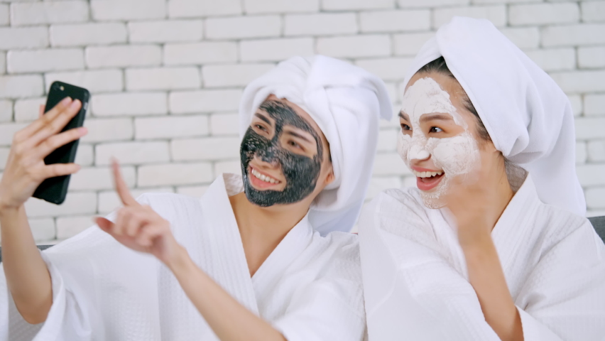 Two happy Asian girls in white bathrobes with facial mask taking photo on smartphone together in living room.