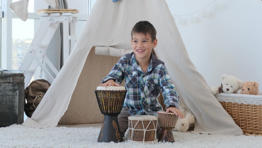 Little boy playing on ethnic drums djembe in room with wigwam