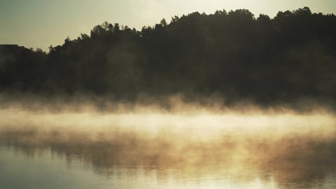 Fog on lake. Golden sunlight rising moving orange mist on water surface in autumn. Forest background and beautiful peaceful reflections 