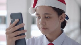 Slow motion of a smiling high school girl in white uniform student and wearing Santa hat is greeting by facial expression and waving his hand on video call Christmas celebration happily .