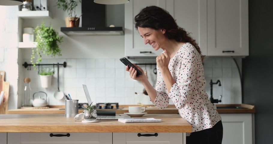 Woman standing in home kitchen hold device receive incredible news on smartphone feels very happy and overjoyed, celebrate online lottery gambling win, great opportunity, good notice from bank concept | Shutterstock HD Video #1060757527