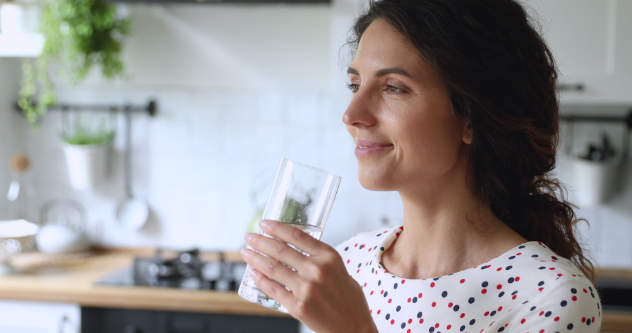 Thirsty smiling 35s woman standing alone in domestic kitchen start new day with healthy life habit, holding glass drinking clean mineral natural still water close up view. Lifestyle healthcare concept | Shutterstock HD Video #1060757560