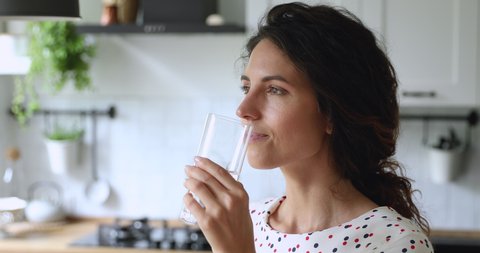 Thirsty smiling 35s woman standing alone in domestic kitchen start new day with healthy life habit, holding glass drinking clean mineral natural still water close up view. Lifestyle healthcare concept