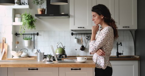 Woman standing in home cozy kitchen use laptop do freelance work looking focused deep lost in thoughts, thinking solving issues distantly makes research, author writing creative work online concept
