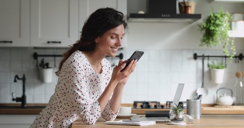 Smiling millennial 35s woman standing in kitchen holding mobile phone looks at screen scroll photos in social media network, spend time on internet websites, choose goods in fashion e-stores at home.