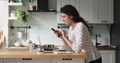 Woman resting standing in kitchen lean at dining table hold smartphone choose goods on internet, make food delivery order on-line. Modern wireless tech simplifies life, easy e services usage concept