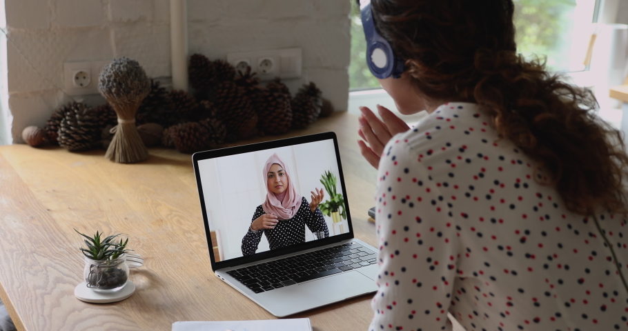 Laptop screen view over female shoulder during videocall communication. Two multicultural women indian ethnicity businesswoman wear hijab talk with caucasian colleague in headphones use video call app Royalty-Free Stock Footage #1060757614